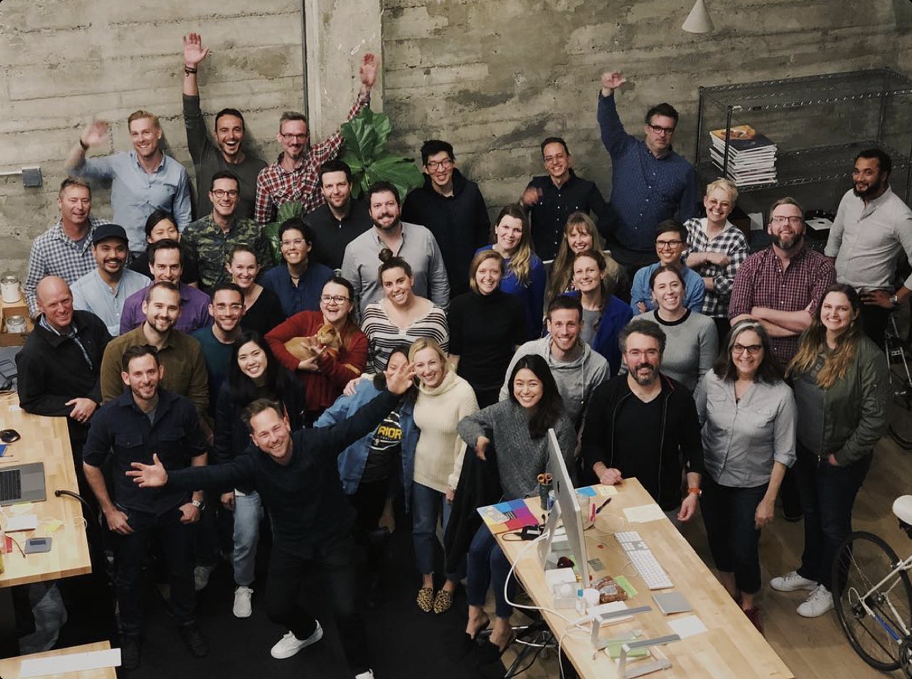 Large team group photo in a concrete-walled studio, everyone waving